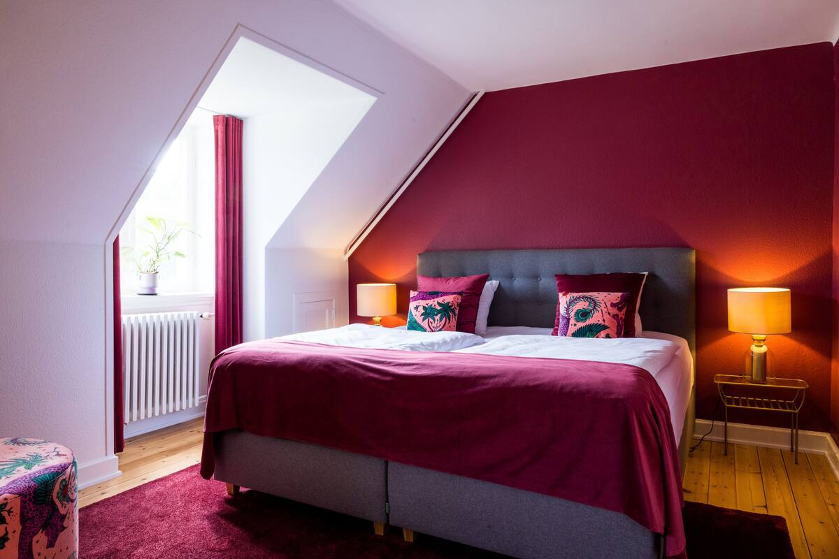 A bedroom with a sloped ceiling, a bed with a grey headboard, two bedside lamps, and a window with red curtains.
