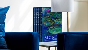 A room with a blue sofa, a small table, a lamp, and a Monet book on the wall. - Gem Suites Holiday Apartments (Augustenborg)