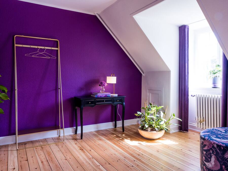 Una habitación con paredes moradas, suelo de madera, una mesa negra con lámpara, un perchero y una ventana con cortinas.