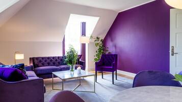 A modern living room with a purple color scheme, featuring a sofa, armchairs, a coffee table, and a lamp.