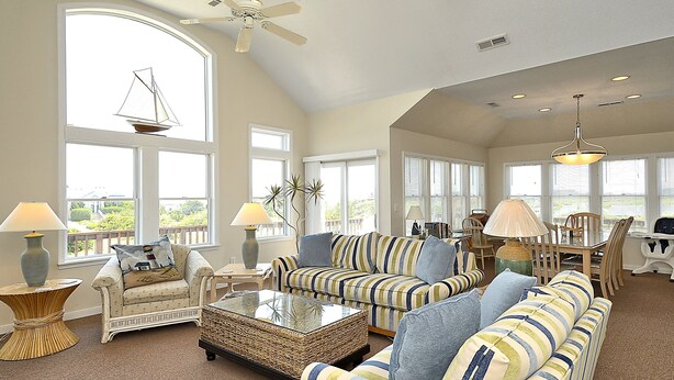 A spacious living room with large windows, a ceiling fan, and a dining area with chairs and a table. - 4668- 85 Ocean Blvd, Walk to Beach | Private Pool & Game Room (Southern Shores)
