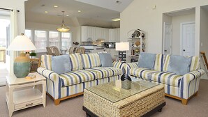 A spacious living room with a striped sofa, a glass coffee table, and a wicker ottoman. - 4668- 85 Ocean Blvd, Walk to Beach | Private Pool & Game Room (Southern Shores)