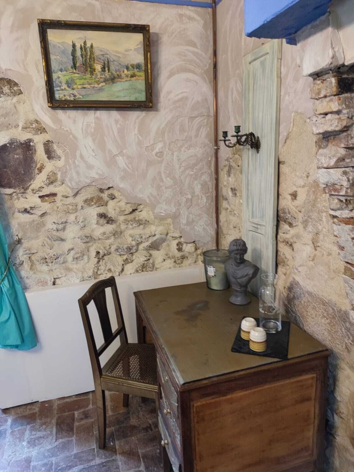 A rustic room with a stone wall, a wooden table, a chair, a bust sculpture, and a framed painting.