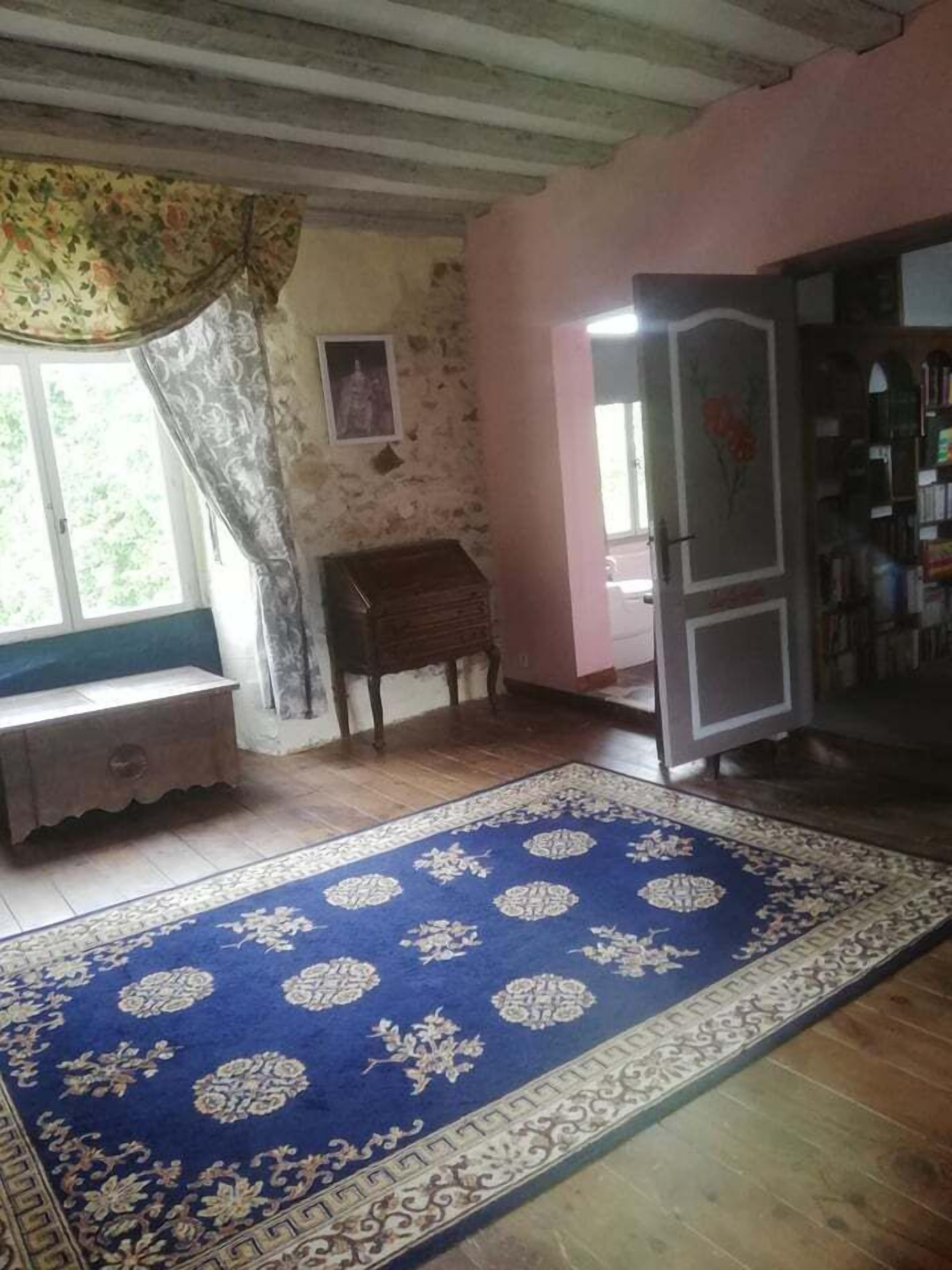Une pièce avec un plancher en bois, un tapis à motifs bleus, un banc en bois, une petite table, une bibliothèque et une fenêtre avec un rideau.