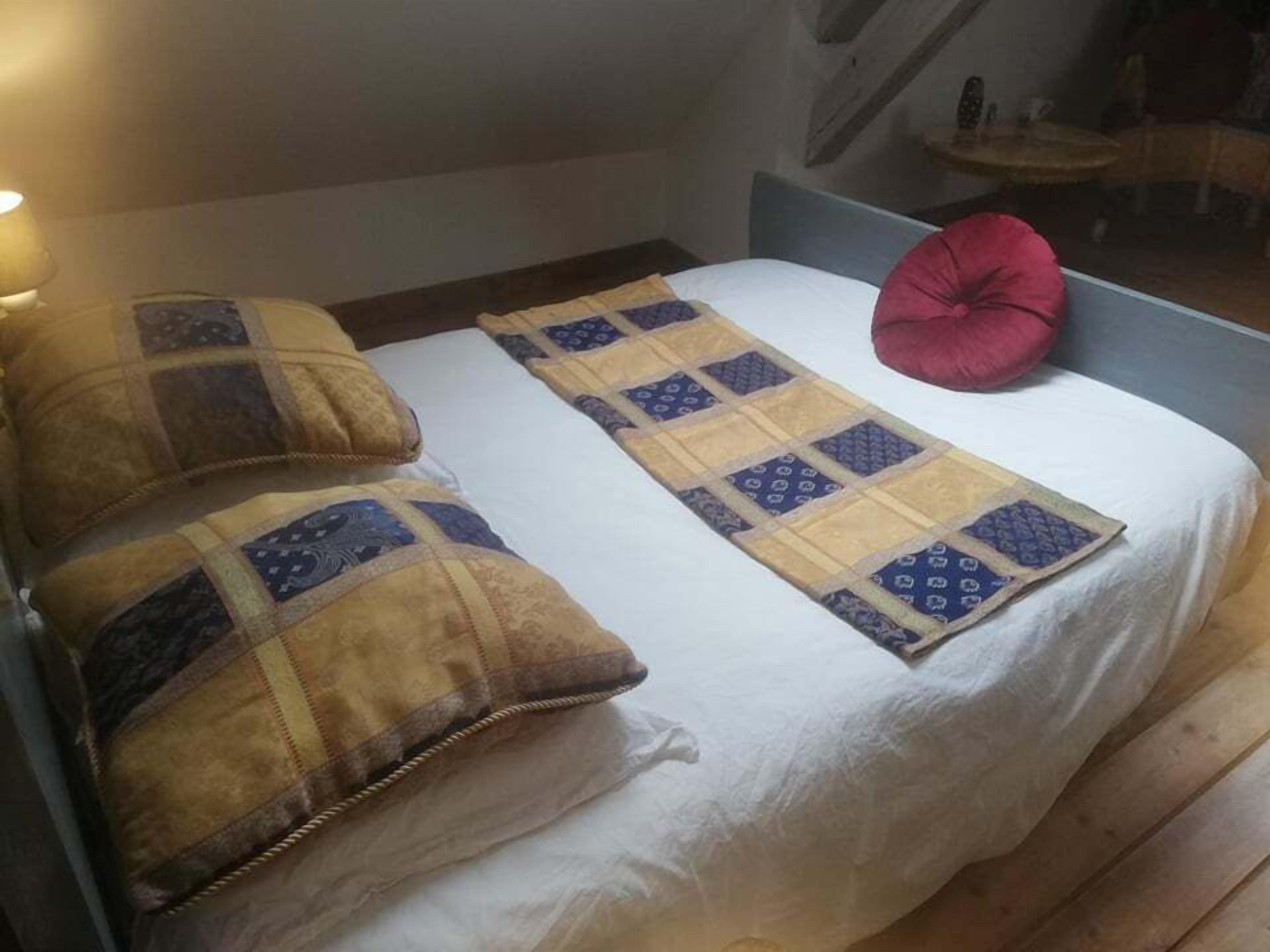 A bed with a white mattress, two pillows, a quilt, and a red accent pillow.
