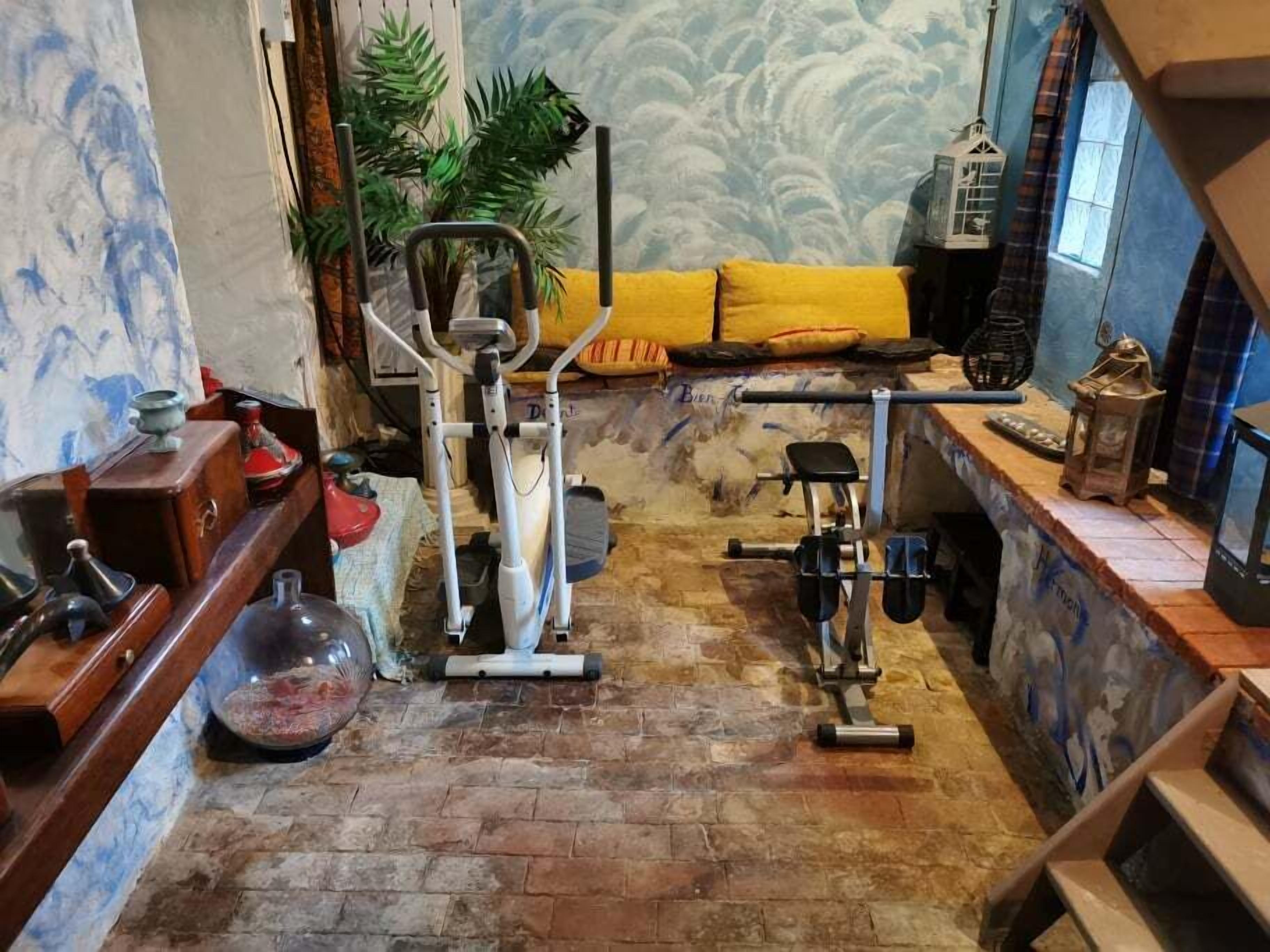 Une pièce avec un sol en briques, du matériel d’exercice, un canapé jaune et une fenêtre avec des rideaux.