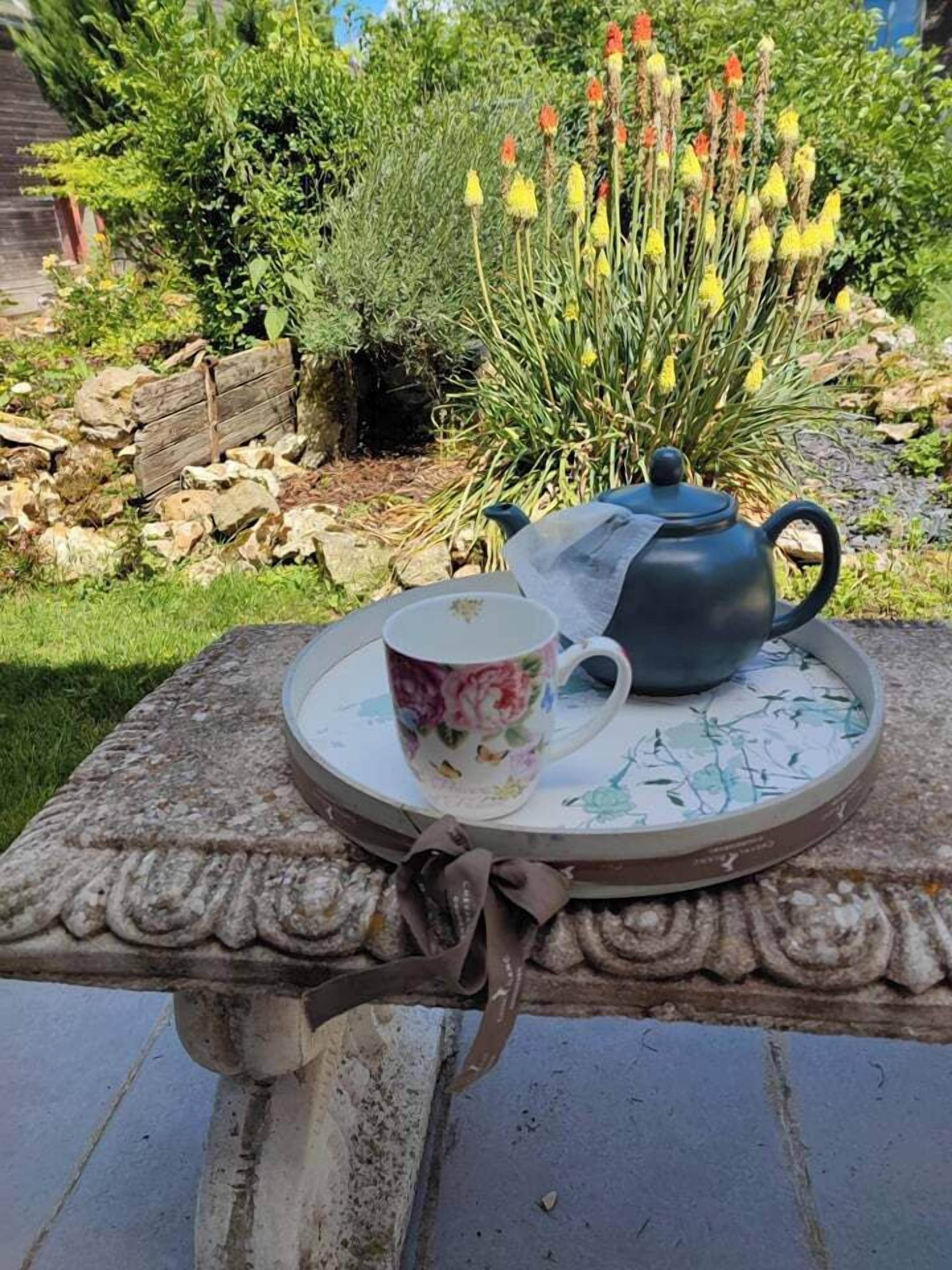 Ein Tisch mit Teekanne und bemalter Tasse auf einer verzierten Ablage im Garten.