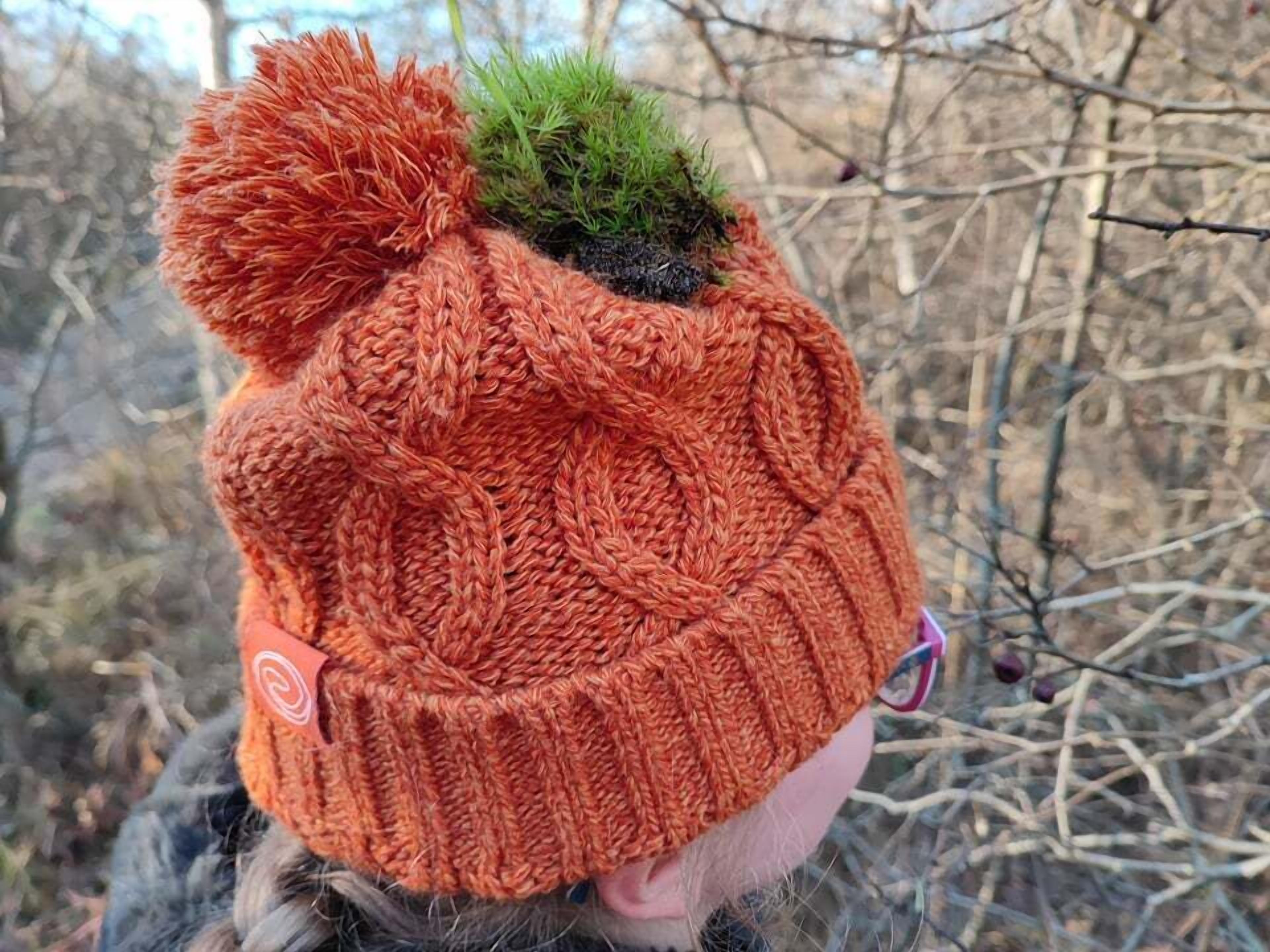 A knitted hat with a pom-pom and a small green moss-like decoration.