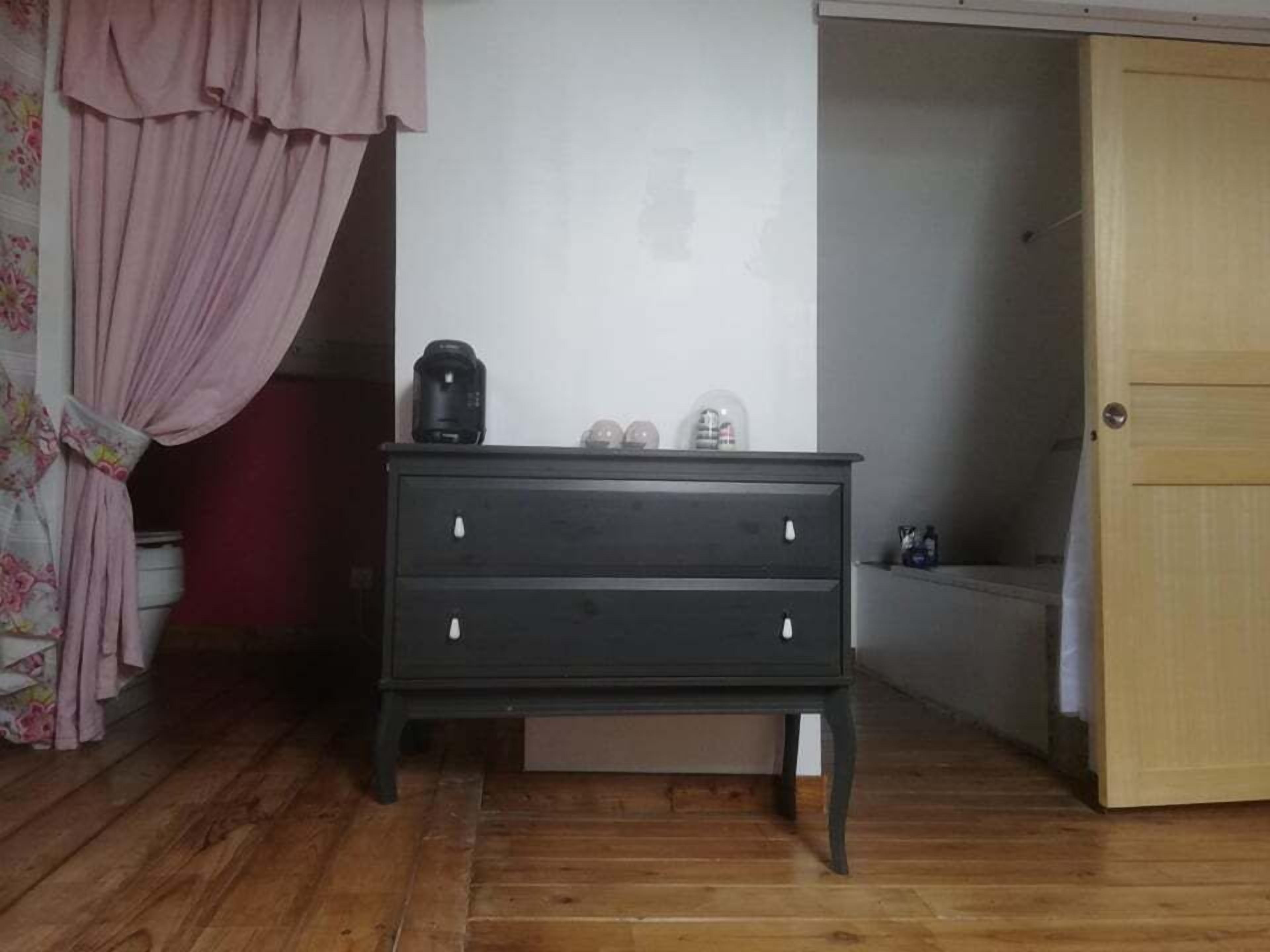 Una habitación pequeña con piso de madera, un aparador oscuro de tres cajones, una cortina rosa y una puerta de madera.
