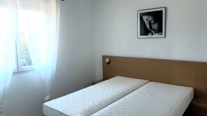 Une pièce simple, avec un lit, une fenêtre ornée de rideaux légers et un tableau encadré accroché au mur.