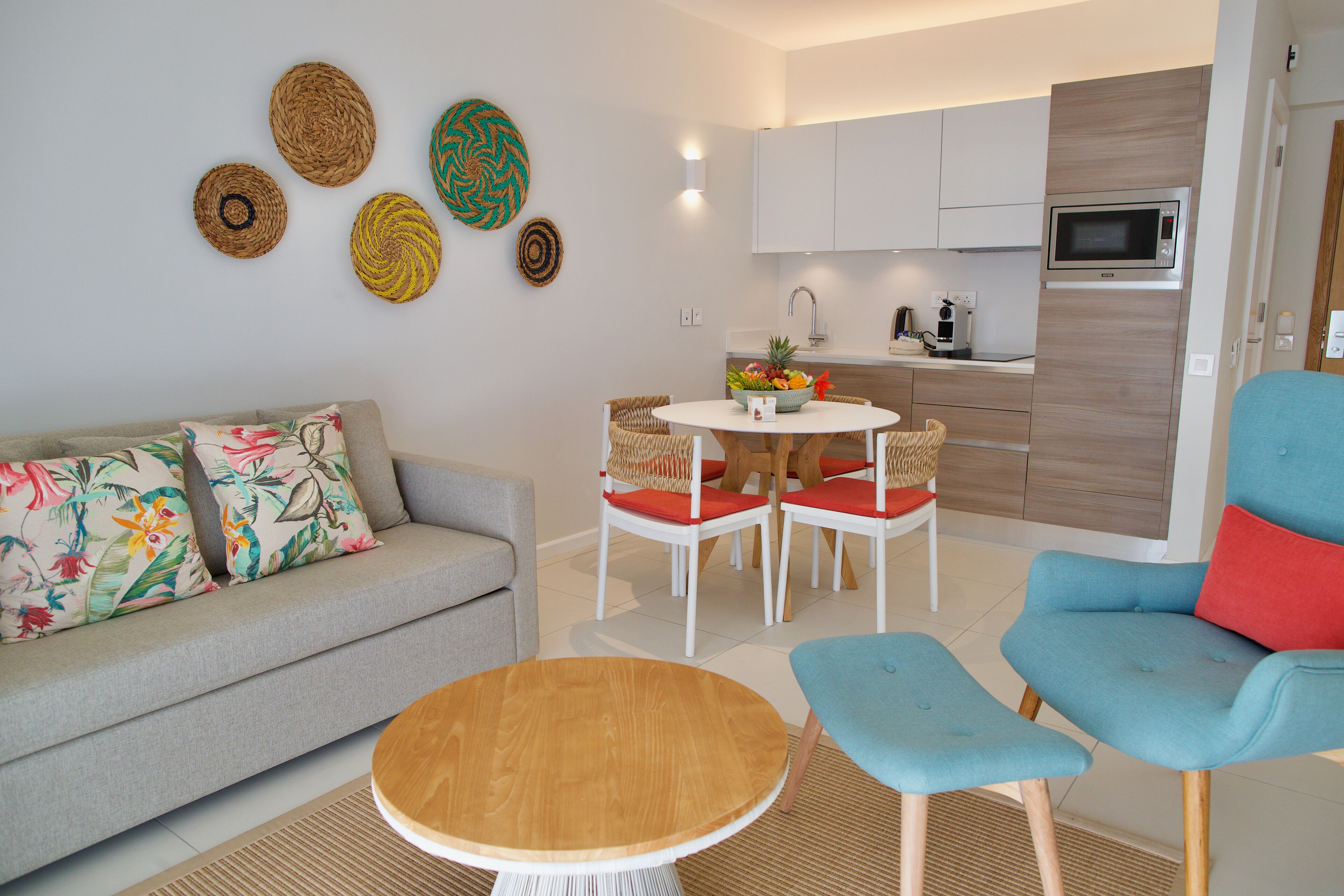 Un salon moderne avec un canapé, une table basse ronde et un fauteuil bleu. On y trouve également un coin cuisine avec des meubles blancs et un four à micro-ondes.