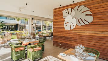 Un comedor con sillas y mesas de mimbre, una pared de madera con un diseño de hojas, y vista al jardín exterior.