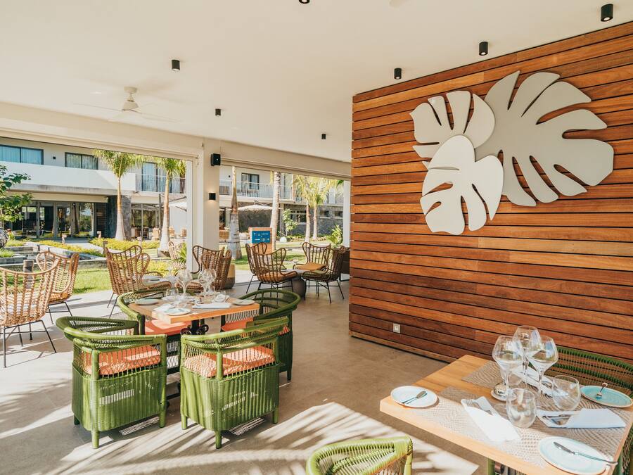 Un comedor con sillas y mesas de mimbre, una pared de madera con un diseño de hojas, y vista al jardín exterior.