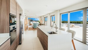 A modern kitchen with a central island, a dining area with a view, and a large sliding glass door leading to a balcony. - Domaine de Grand Baie (Mont Choisy)