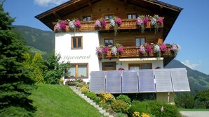 Een houten chalet met twee verdiepingen, met balkons versierd met bloemen, zonnepanelen en een bordje met de tekst 'Alpenrast'.