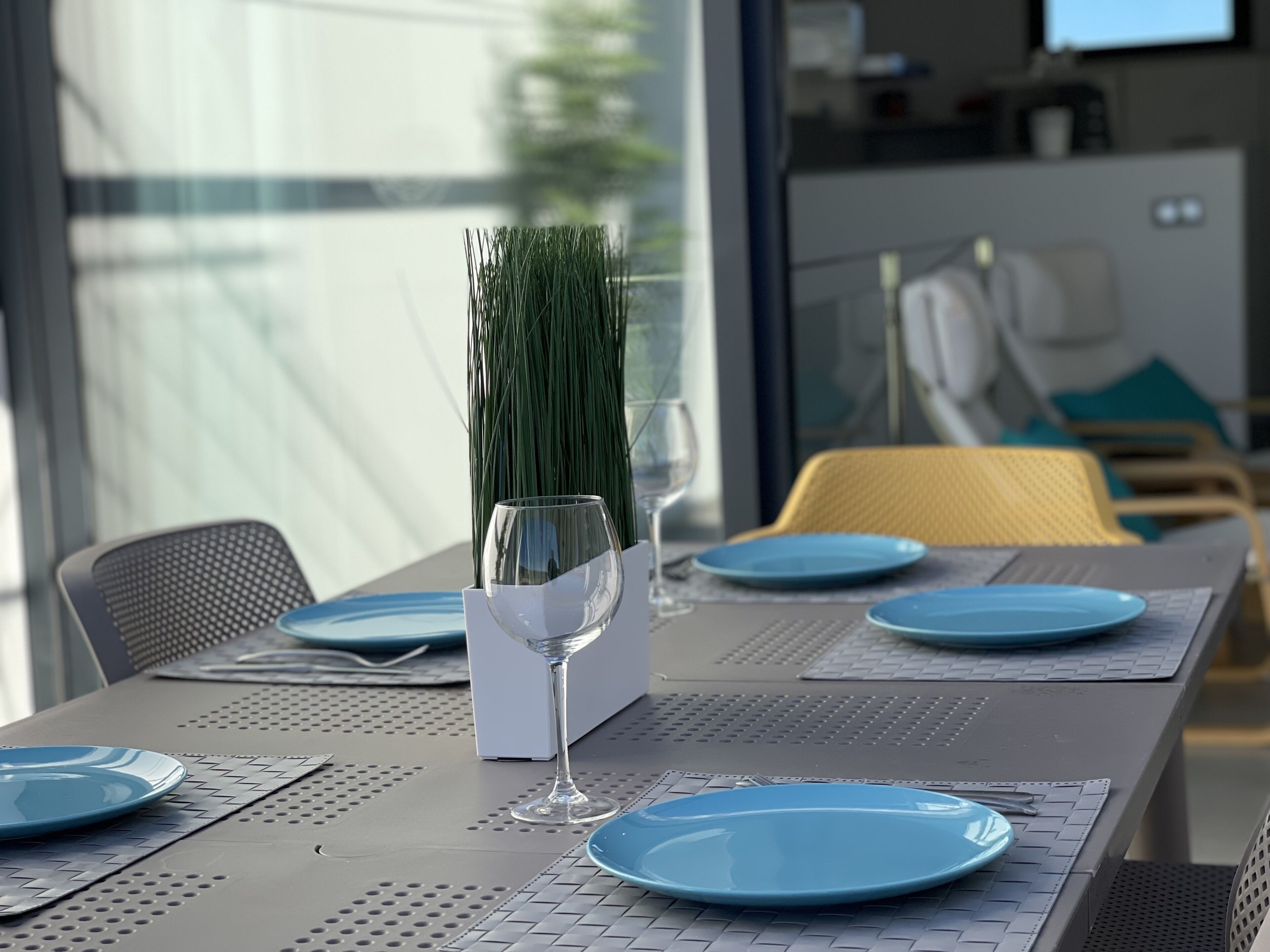 A dining table set with blue plates, a clear wine glass, and a white vase with green plants.