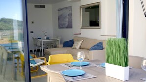 En moderne stue med en glasdør, der fører til en balkon, en sofa med blå puder, et spisebord dækket med blå tallerkener og glas, samt en potteplante.