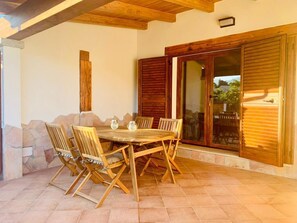 A covered patio with a wooden table and chairs, stone walls, and a sliding glass door. - I Gerani 4 San Teodoro Centro (San Teodoro)