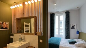 Un bagno con un mobiletto in legno, un lavandino e uno specchio. Nella stanza adiacente, un letto con coperta blu e un cuscino.