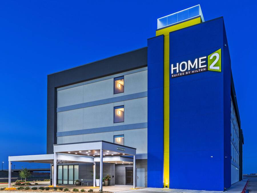 Un moderno edificio de hotel con un letrero destacado que dice 'HOME 2', que presenta una entrada cubierta y un cielo despejado al fondo.