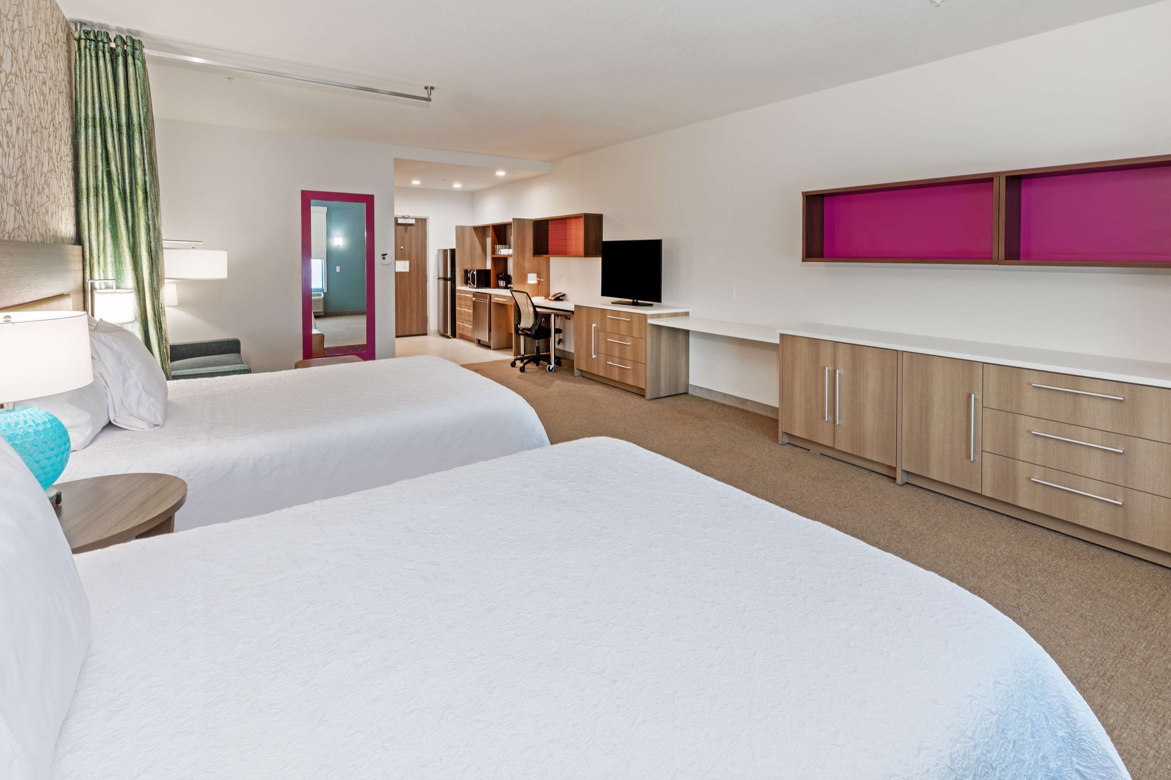 A hotel room with two beds, a desk, a kitchenette, and a TV.