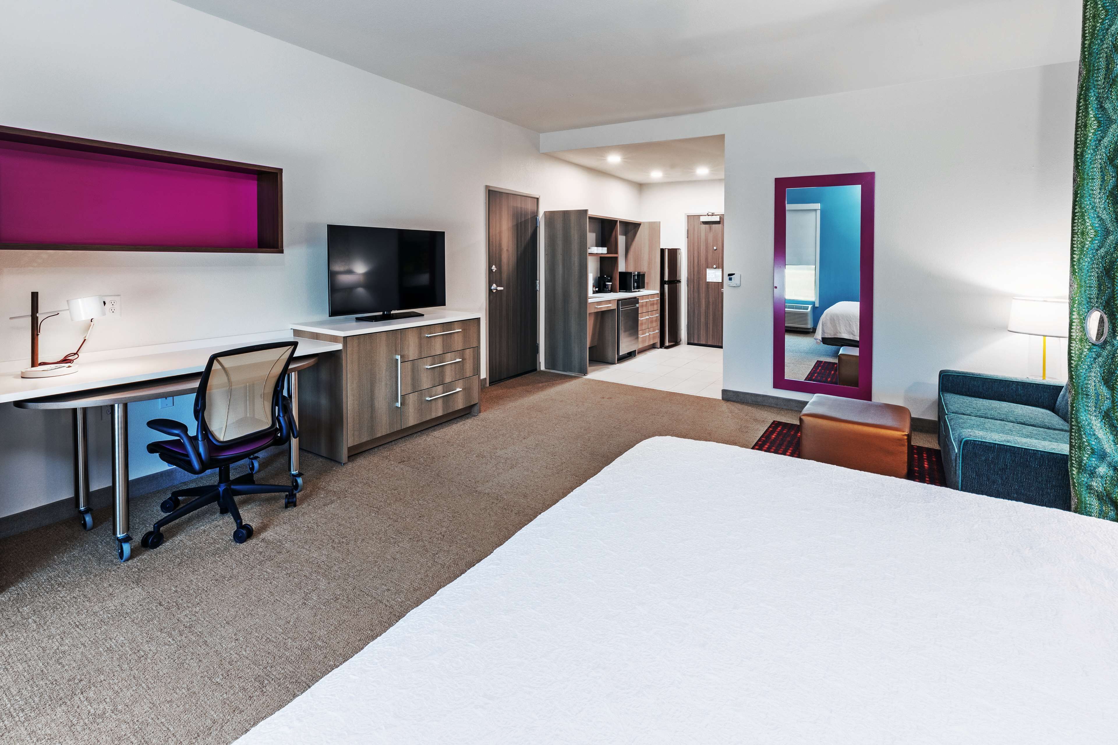 Una habitación de hotel moderna con una cama grande, un escritorio con una silla, un televisor, una kitchenette y un baño visible a través de un espejo.