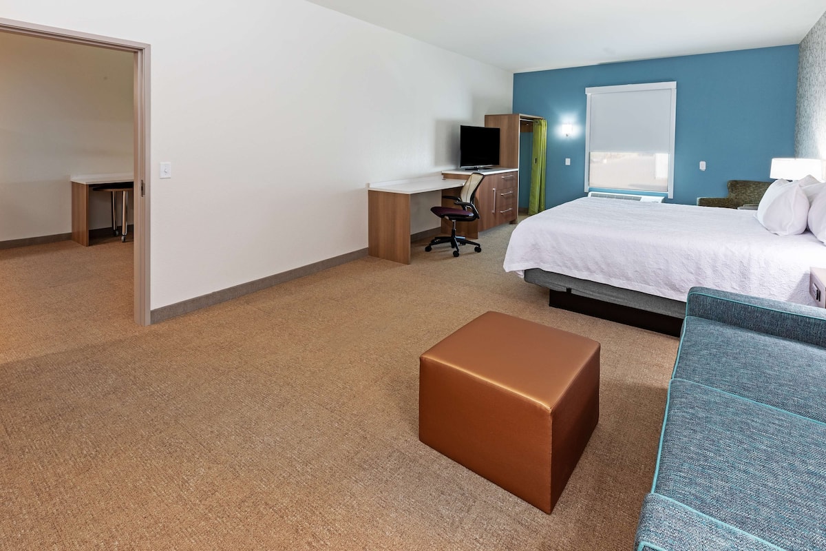 A hotel room with a bed, a desk with a computer, a chair, a brown ottoman, and a blue sofa.