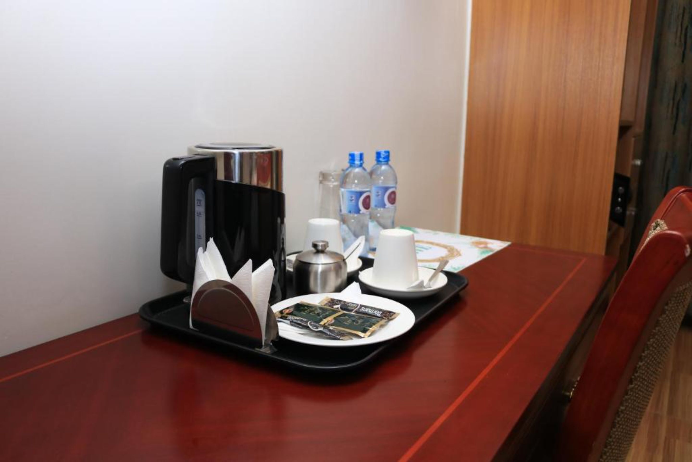A hotel room with a wooden dining table set with a tray containing a coffee maker, two bottles of water, a white cup, a saucer, and a plate with a menu.
