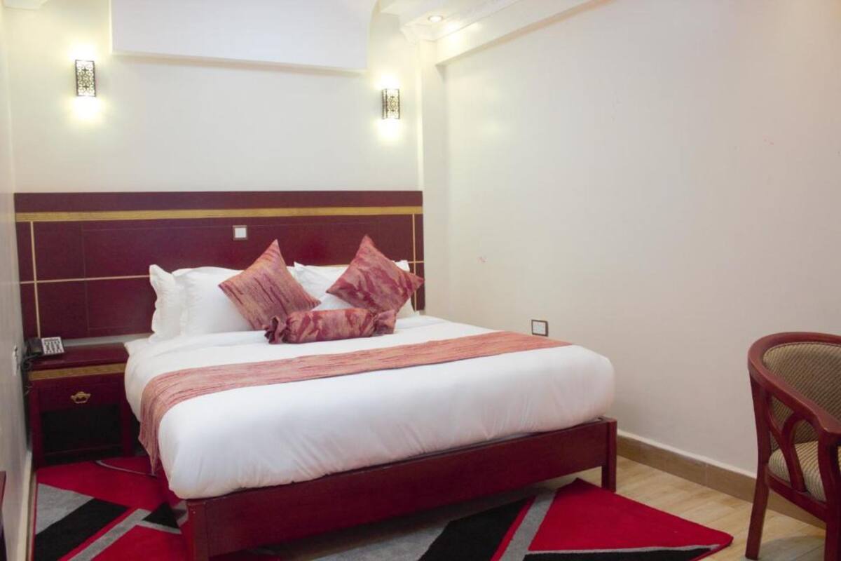 A neatly made bed with a wooden headboard, a red and black rug, and a wooden nightstand with a lamp.
