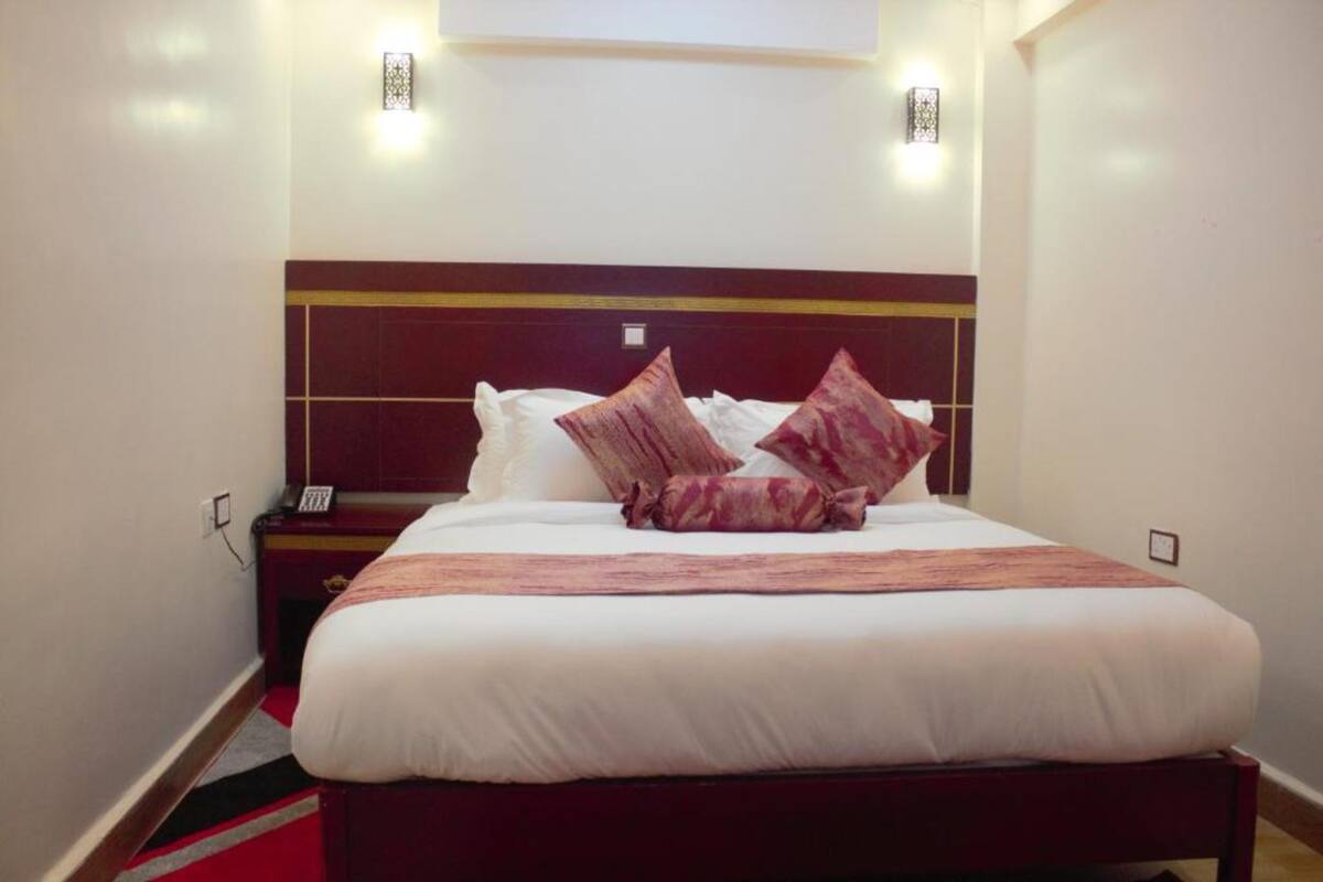 A hotel room with a large bed, a wooden headboard, a bedside table with a lamp, and a red carpet.
