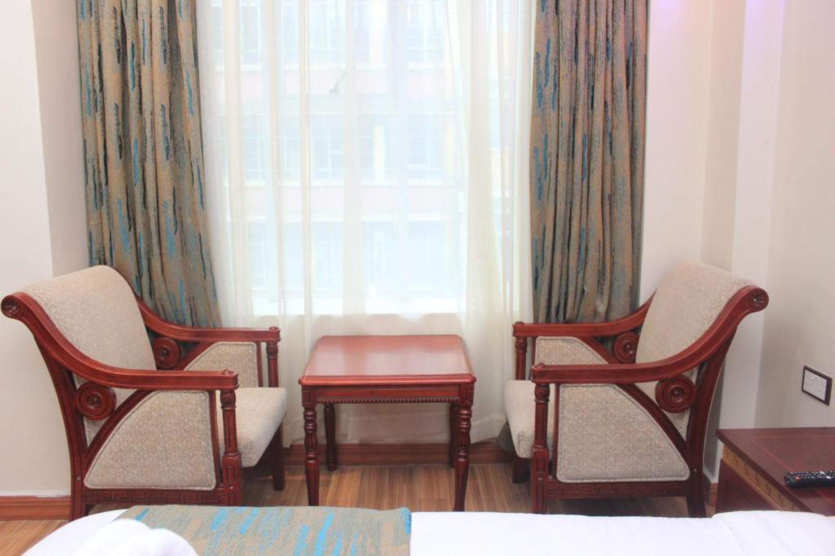 A hotel room with two wooden armchairs, a small wooden table, and a bed with a blue-striped throw.