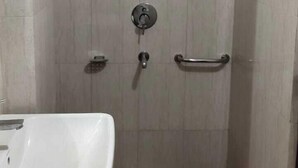 A small, tiled shower stall with a handheld showerhead, a toilet, and a red stool. - Green Leaf (Jhansi)
