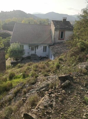 A rustic house with a tiled roof, situated on a hillside with a view of distant mountains.