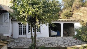 A white building with a tiled patio, a tree, and a covered area with a table and chairs. - Family winegrower's house (Roquebrun)
