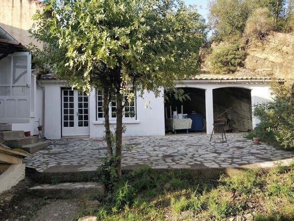 A white building with a tiled patio, a tree, and a covered area with a table and chairs.
