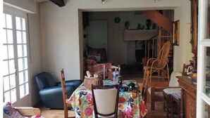A dining area with a table covered in a floral tablecloth, surrounded by wooden chairs. There is a blue sofa and a striped armchair in the foreground. The room has a tiled floor and exposed wooden beams on the ceiling. - Family winegrower's house (Roquebrun)