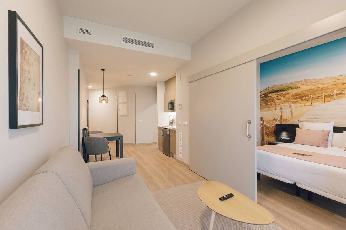 A modern hotel room with a large bed, a sofa, a small table, and a kitchenette.