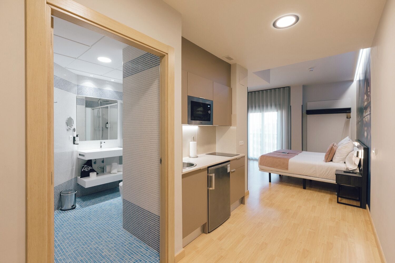 Habitación de hotel compacta con kitchenette, una cama con cabecera, una mesita de noche, un espejo y zona de ducha.
