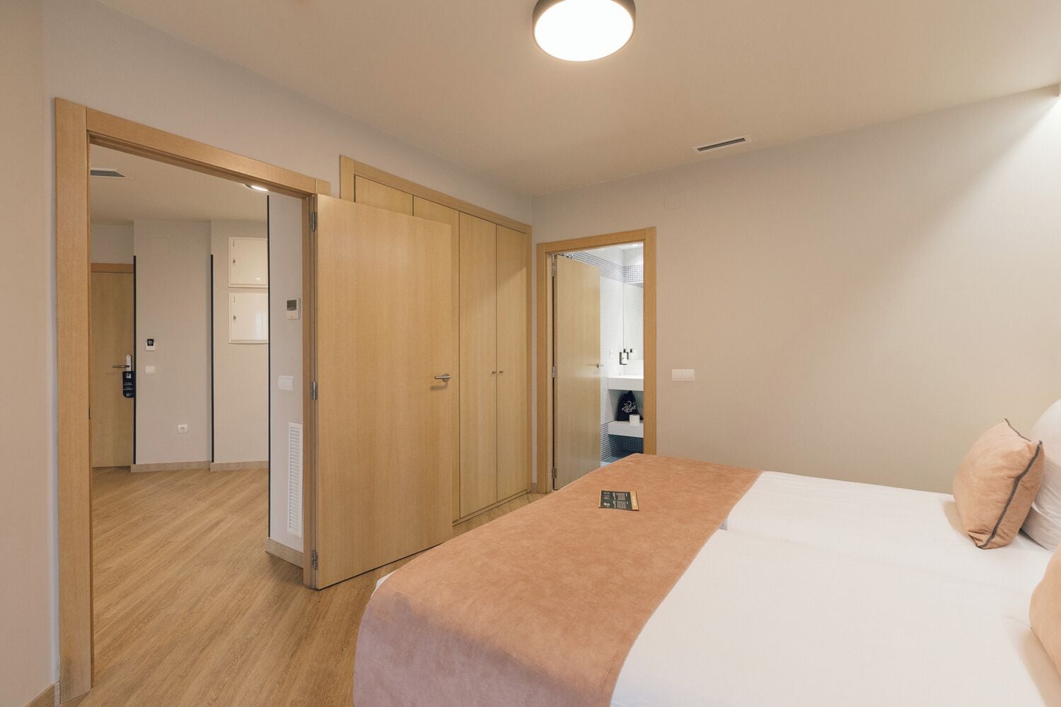 A hotel room with a bed, a wooden wardrobe, and a bathroom visible through a glass door.