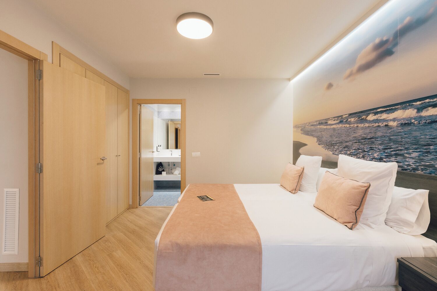 A bedroom with a bed, pillows, a nightstand, a wall-mounted TV, and a view of the ocean through a window.