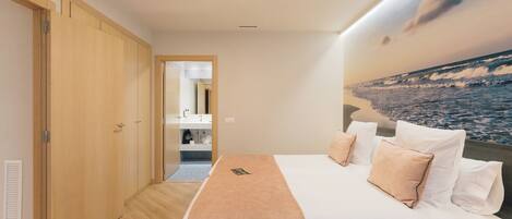 A bedroom with a bed, pillows, a nightstand, a wall-mounted TV, and a view of the ocean through a window.