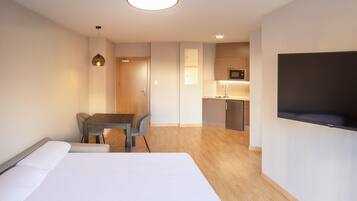Una habitación de hotel moderna con una cama, una mesa de comedor con sillas, una kitchenette y un televisor montado en la pared.