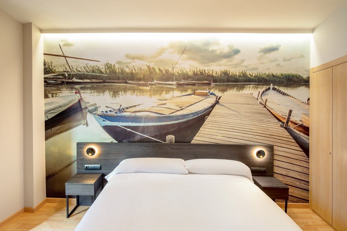 Un dormitorio con un gran mural de un muelle y barcos, un cabecero de madera y una mesita de noche con una lámpara.