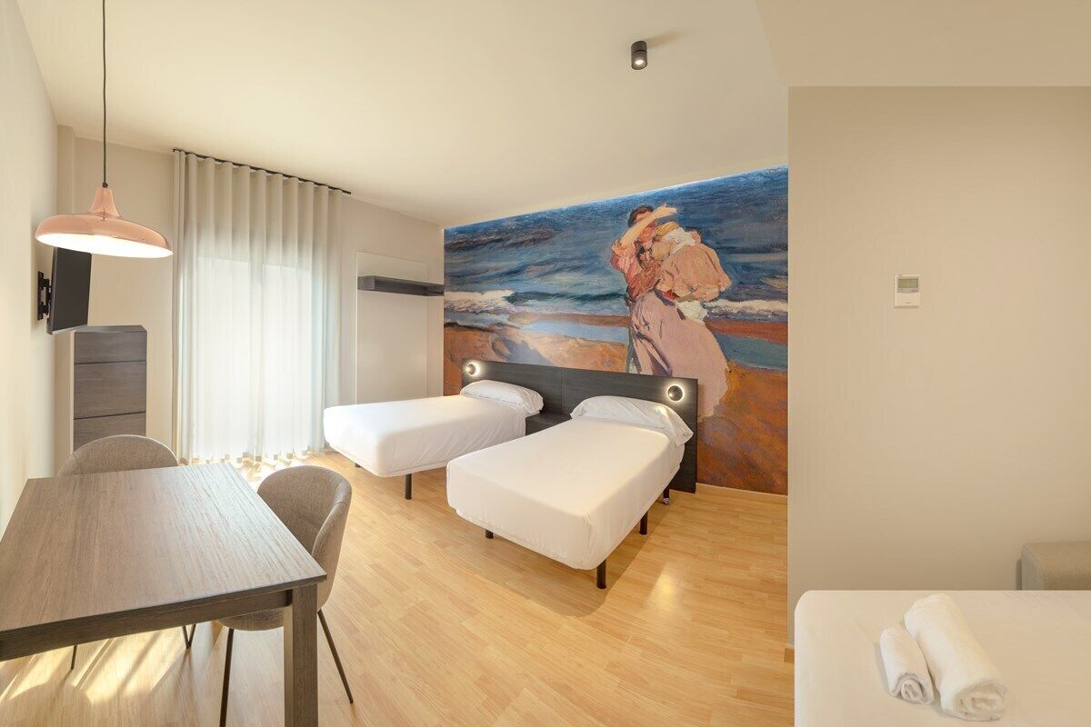 A hotel room with two single beds, a wooden table, a desk, a painting of a woman embracing a child, and a view of the sea.
