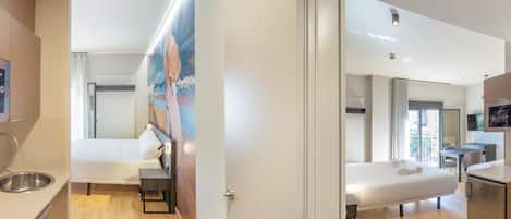 A modern hotel room with a bed, a desk, a kitchenette, and a bathroom.