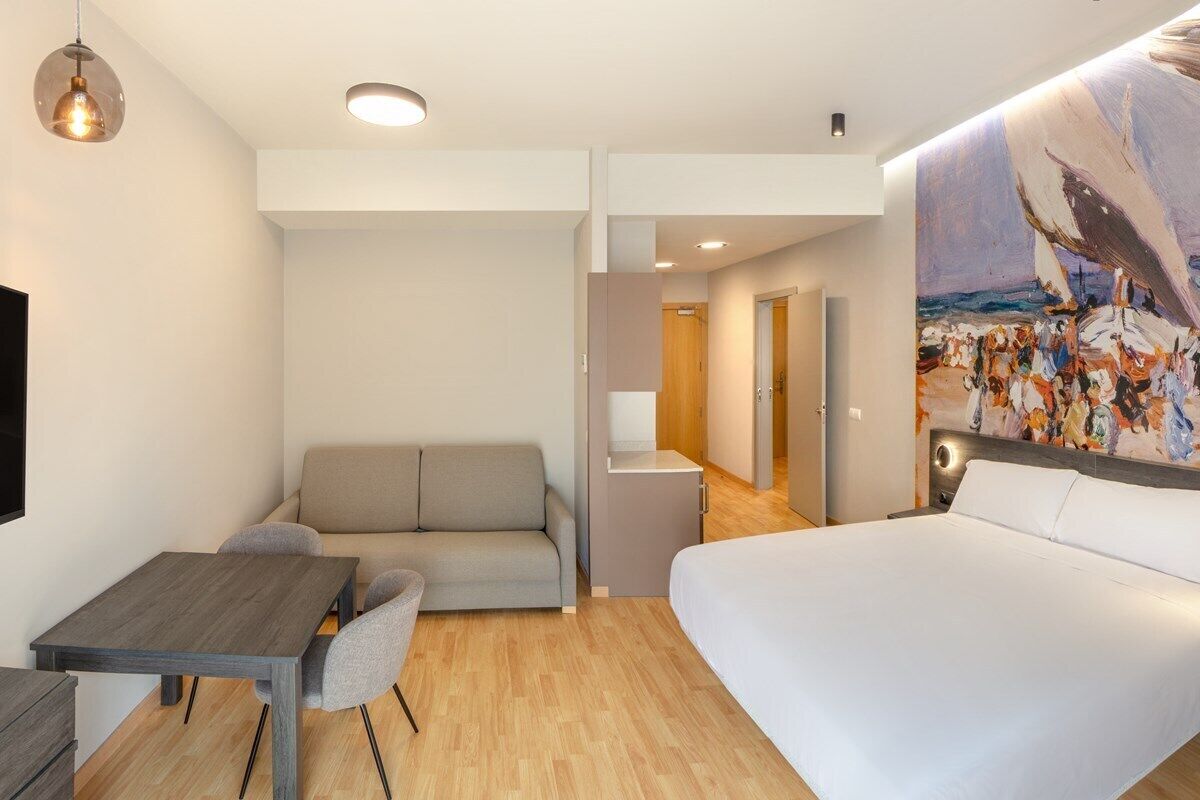 A modern hotel room with a bed, a sofa, a dining table, and a painting on the wall.