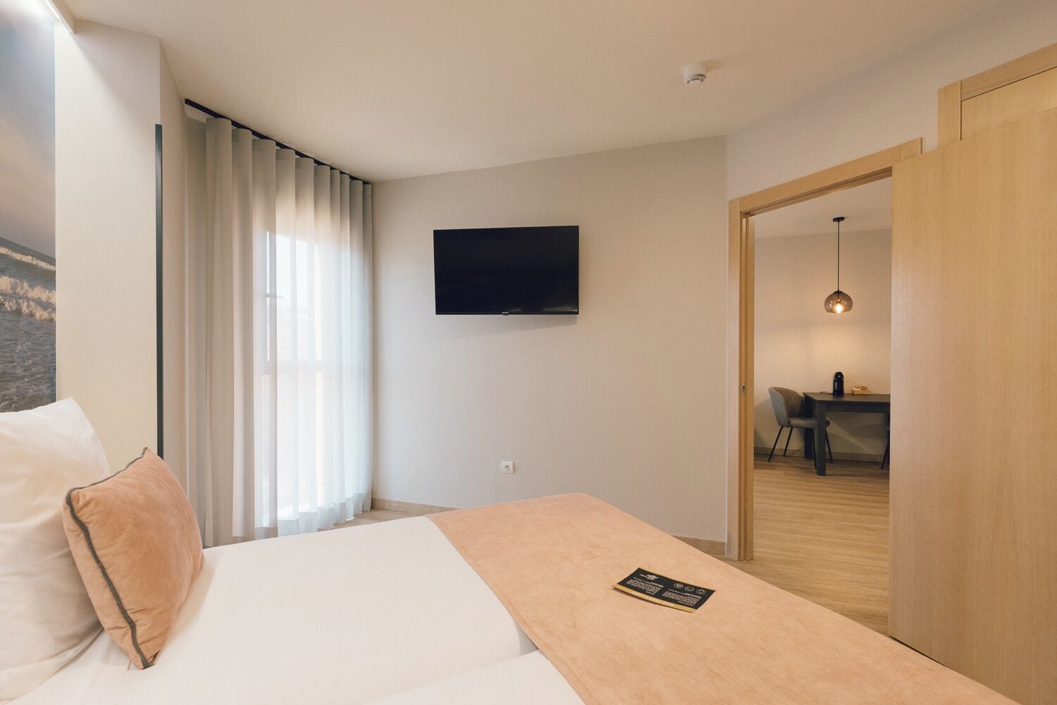 Een hotelkamer met een bed, een aan de muur bevestigde televisie, een bureau met een stoel en een eethoek zichtbaar door een open deur.