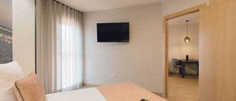 Une chambre d’hôtel avec un lit, un téléviseur fixé au mur, un bureau avec une chaise et un coin repas visible à travers une porte ouverte.