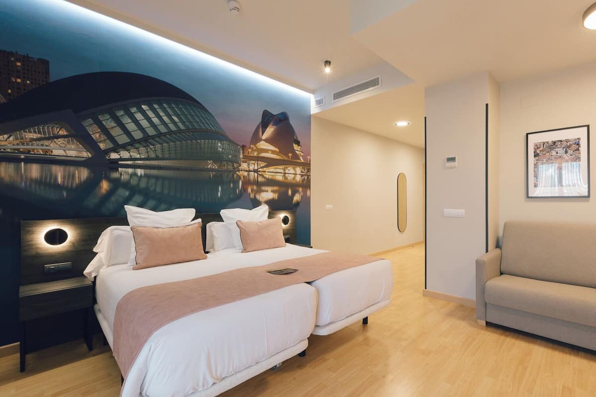 A modern hotel room with a large bed, a sofa, and a mural of a cityscape on the wall.