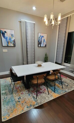 A modern dining room with a white table, black chairs, a colorful rug, abstract paintings, and a chandelier.